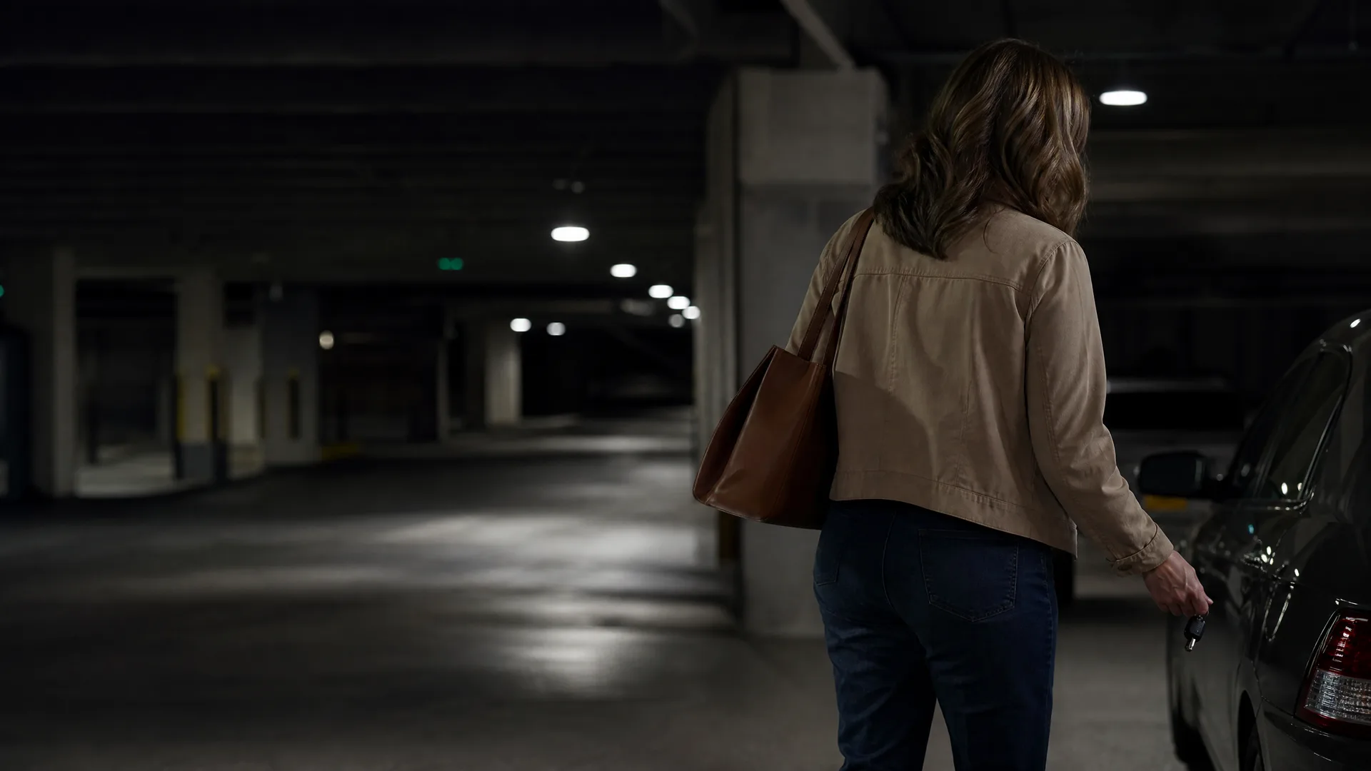 A woman seen from behind in a tan jacket and jeans with a brown shoulder bag walks through a dim concrete parking garage holding a key fob near a parked car.
